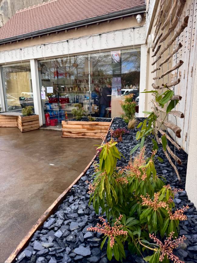 Aménagement d'entrée d'un magasin, bacs en bois, rempli avec des pierres noires et végétations.