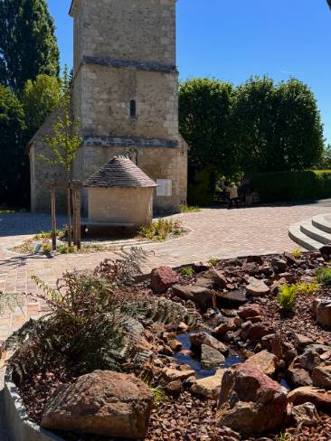 Une allée d'entrée de maison individuelle avec un jardiné devant rempli de pierres, de fougères et d'eau. Au fond une petite église.